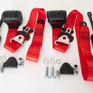May include: Three red safety harnesses with black buckles and metal clips. The harnesses are attached to black metal buckles with metal clips. The harnesses are on a white surface.