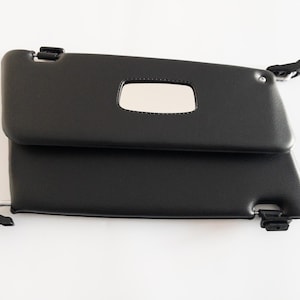 May include: Two black car sun visors with integrated mirrors. The visors are rectangular with rounded corners and feature mounting hardware. The mirrors are rectangular with rounded corners. The visors are shown against a white background.