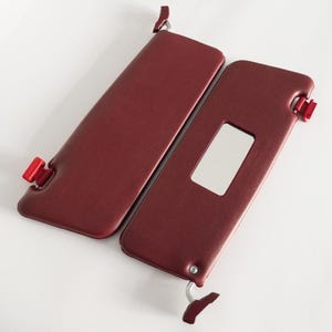 May include: Two burgundy car sun visors with red accents. One visor has a rectangular mirror. The visors are made of a smooth material and are designed to block sunlight. Automotive accessory.