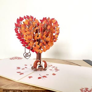 May include: A pop-up card with a heart-shaped tree in autumnal colours. Two small hedgehogs sit beneath the tree. A heart with the word "Love" hangs from the tree. The card is open and has a light beige colour. The card is made of paper.