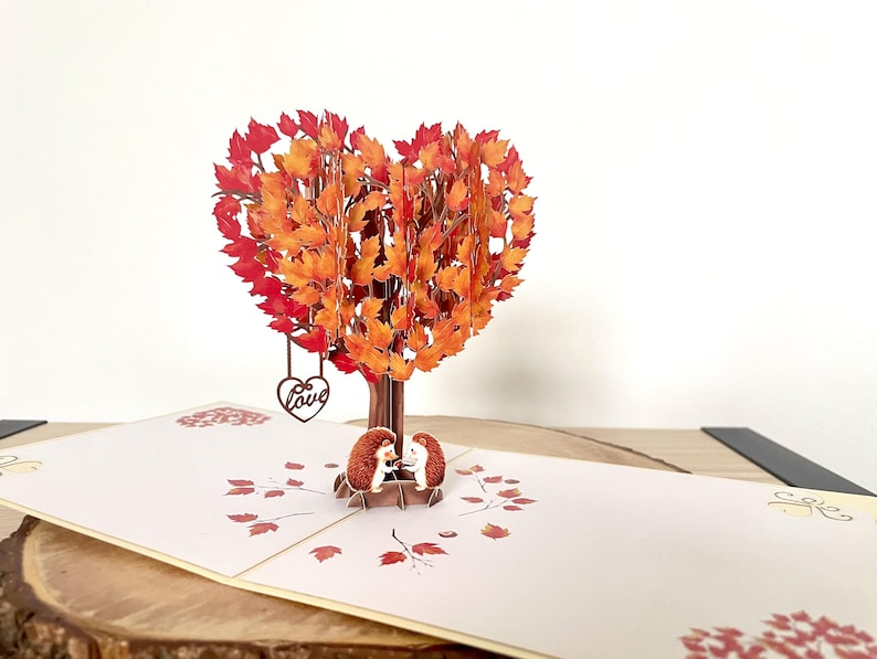 May include: A pop-up card with a heart-shaped tree in autumnal colours. Two small hedgehogs sit beneath the tree. A heart with the word "Love" hangs from the tree. The card is open and has a light beige colour. The card is made of paper.