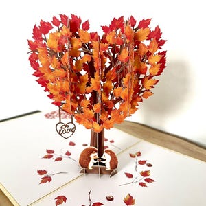 May include: A 3D pop-up card with a heart-shaped tree, its leaves in shades of orange and red. Two hedgehogs are at the base. A small heart with the word "Love" hangs from the tree, evoking an autumnal theme.