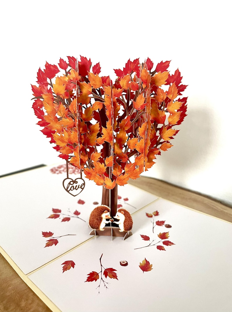 May include: A 3D pop-up card with a heart-shaped tree, its leaves in shades of orange and red. Two hedgehogs are at the base. A small heart with the word "Love" hangs from the tree, evoking an autumnal theme.