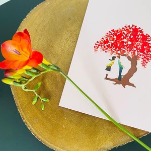 K&ouml;nnte beinhalten: Eine rosa Karte mit einem roten Baum und zwei Personen, die darunter stehen. Die Karte liegt auf einer Holzoberfl&auml;che mit einer einzelnen orangefarbenen Blume.