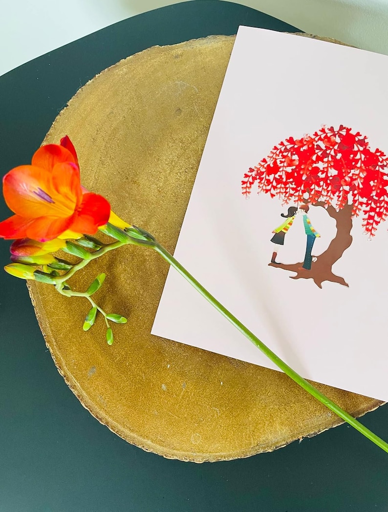 K&ouml;nnte beinhalten: Eine rosa Karte mit einem roten Baum und zwei Personen, die darunter stehen. Die Karte liegt auf einer Holzoberfl&auml;che mit einer einzelnen orangefarbenen Blume.