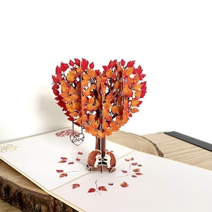 May include: A pop-up card with a heart-shaped tree made of orange and red leaves. Two hedgehogs are under the tree. The card is on a white surface with printed leaves and a wooden base.
