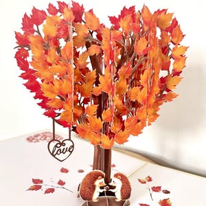 May include: A pop-up greeting card with a heart-shaped tree, its leaves in shades of orange and red. A brown heart ornament with the word "Love" dangles from the tree. Two hedgehogs are at the base of the tree.