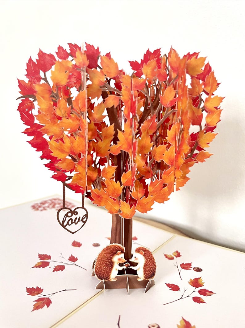 May include: A pop-up greeting card with a heart-shaped tree, its leaves in shades of orange and red. A brown heart ornament with the word "Love" dangles from the tree. Two hedgehogs are at the base of the tree.