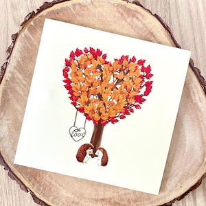 May include: A square greeting card with a heart-shaped tree in autumn colours. Two hedgehogs stand beneath the tree, and a swing with the word "Love" hangs from a branch. The card rests on a wooden surface.