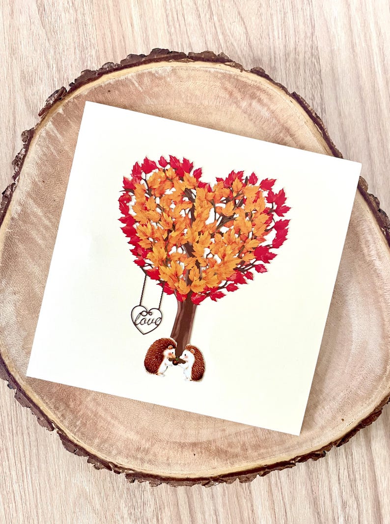 May include: A square greeting card with a heart-shaped tree in autumn colours. Two hedgehogs stand beneath the tree, and a swing with the word "Love" hangs from a branch. The card rests on a wooden surface.