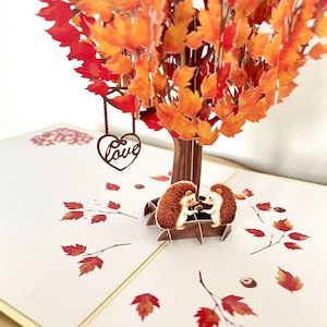 May include: A pop-up greeting card depicting an autumnal tree with orange and red leaves. Two hedgehogs sit on a bench. A brown heart with the word "Love" hangs from the tree. Scattered leaves and nuts complete the design.