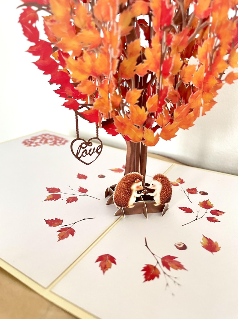 May include: A pop-up greeting card depicting an autumnal tree with orange and red leaves. Two hedgehogs sit on a bench. A brown heart with the word "Love" hangs from the tree. Scattered leaves and nuts complete the design.