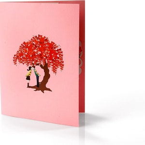 K&ouml;nnte beinhalten: Eine rosa Pop-up-Karte mit einem 3D-Design eines Paares, das sich unter einem Baum mit roten herzf&ouml;rmigen Bl&auml;ttern k&uuml;sst.