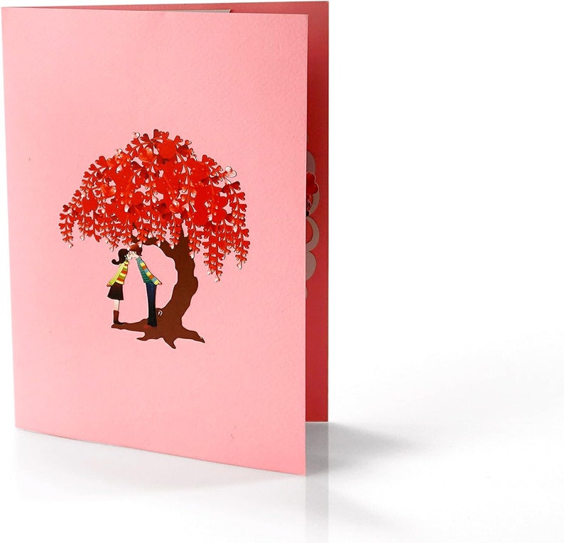K&ouml;nnte beinhalten: Eine rosa Pop-up-Karte mit einem 3D-Design eines Paares, das sich unter einem Baum mit roten herzf&ouml;rmigen Bl&auml;ttern k&uuml;sst.
