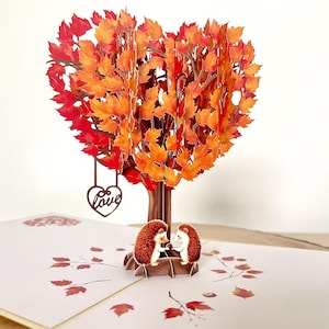 May include: A pop-up card with a heart-shaped tree in autumnal colours. Two hedgehog figures are at the base. A wooden heart with the word "Love" hangs from the tree. The card is open, revealing a detailed design.