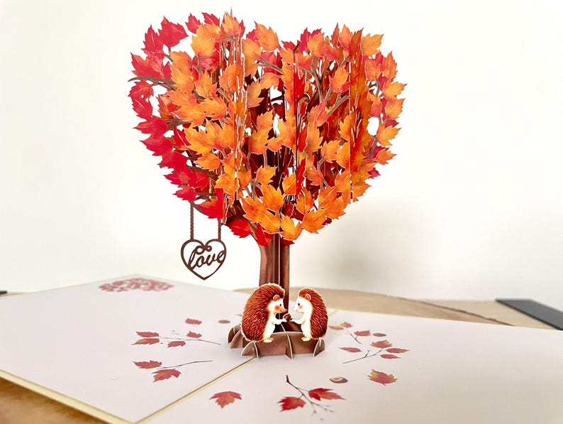 May include: A pop-up card with a heart-shaped tree in autumnal colours. Two hedgehog figures are at the base. A wooden heart with the word "Love" hangs from the tree. The card is open, revealing a detailed design.