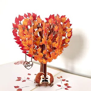 May include: A 3D pop-up card depicting a tree with a heart-shaped crown of red and orange autumn leaves. Two hedgehogs are beneath the tree. A heart pendant with the word "love" hangs from the branches.