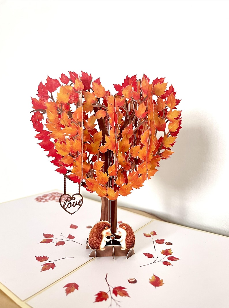 May include: A 3D pop-up card depicting a tree with a heart-shaped crown of red and orange autumn leaves. Two hedgehogs are beneath the tree. A heart pendant with the word "love" hangs from the branches.