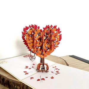 May include: A pop-up card with a heart-shaped tree made of orange and red leaves. Two hedgehogs are under the tree. The card is on a white surface with printed leaves and a wooden base.