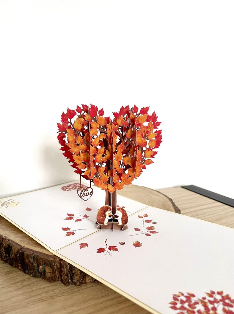 May include: A pop-up card with a heart-shaped tree made of orange and red leaves. Two hedgehogs are under the tree. The card is on a white surface with printed leaves and a wooden base.