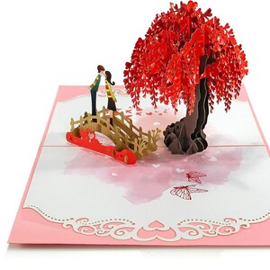 K&ouml;nnte beinhalten: Eine 3D-Pop-up-Karte mit einem Paar, das sich auf einer Br&uuml;cke unter einem roten Trauerweidenbaum k&uuml;sst. Die Karte hat einen rosa Hintergrund mit wei&szlig;en Herzen und Schmetterlingen.