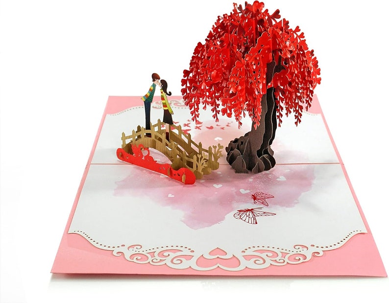K&ouml;nnte beinhalten: Eine 3D-Pop-up-Karte mit einem Paar, das sich auf einer Br&uuml;cke unter einem roten Trauerweidenbaum k&uuml;sst. Die Karte hat einen rosa Hintergrund mit wei&szlig;en Herzen und Schmetterlingen.