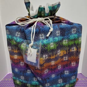 Könnte beinhalten: Ein Geschenkbeutel mit Kordelzug und einem lebendigen Batikmuster in Blau-, Lila-, Grün- und Orangetönen, verziert mit weißen Schneeflocken. Der Beutel ist mit einer weißen Kordel gebunden und enthält ein klares Etikett.