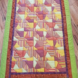 Può includere: Una trapunta patchwork con un motivo geometrico di triangoli in tonalità di arancione, giallo, verde e viola. La trapunta è bordata con un tessuto verde e un tessuto marrone.