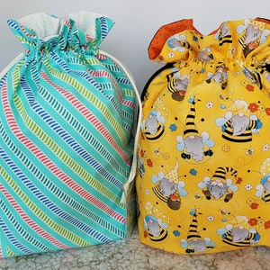 Puede incluir: Dos bolsas de cordón, una con un patrón de rayas azules y verdes y la otra con un fondo amarillo y un patrón de gnomos con trajes de abeja.