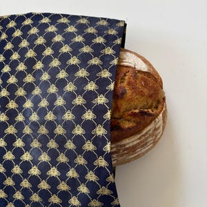 Puede incluir: Una hogaza de pan redonda parcialmente cubierta por una bolsa de pan azul marino. La bolsa presenta un patrón de abejas doradas. El pan tiene una corteza dorada. La bolsa es de tela.