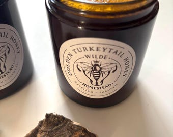 May include: A close-up of two dark brown glass jars filled with golden liquid, labeled "Golden Turkeytail Honey." A dried, multi-toned mushroom sits in front of the jars. The labels feature a bee illustration and the words "Mycelium Turmeric."
