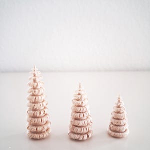 Pode incluir: Três árvores de Natal decorativas rosa claro de tamanhos variados. Cada árvore é construída a partir de elementos circulares empilhados, criando uma aparência texturizada e em camadas. As árvores estão sobre uma superfície branca contra um fundo branco liso.