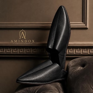 May include: A pair of black leather slip-on shoes with a pointed toe and a textured finish. One shoe is positioned vertically, while the other rests horizontally. The logo AMINDOX HANDCRAFTED IN MOROCCO is visible in gold text on the left side.
