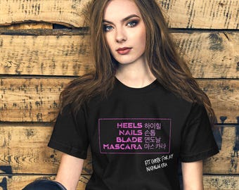May include: A black t-shirt with the words "HEELS, NAILS, BLADE, MASCARA" in a pink rectangle. The text is in both English and Korean. The shirt also has the text "FIT (HECK) FOR AY NIRALA ERA" in white. The person is wearing dark blue jeans.