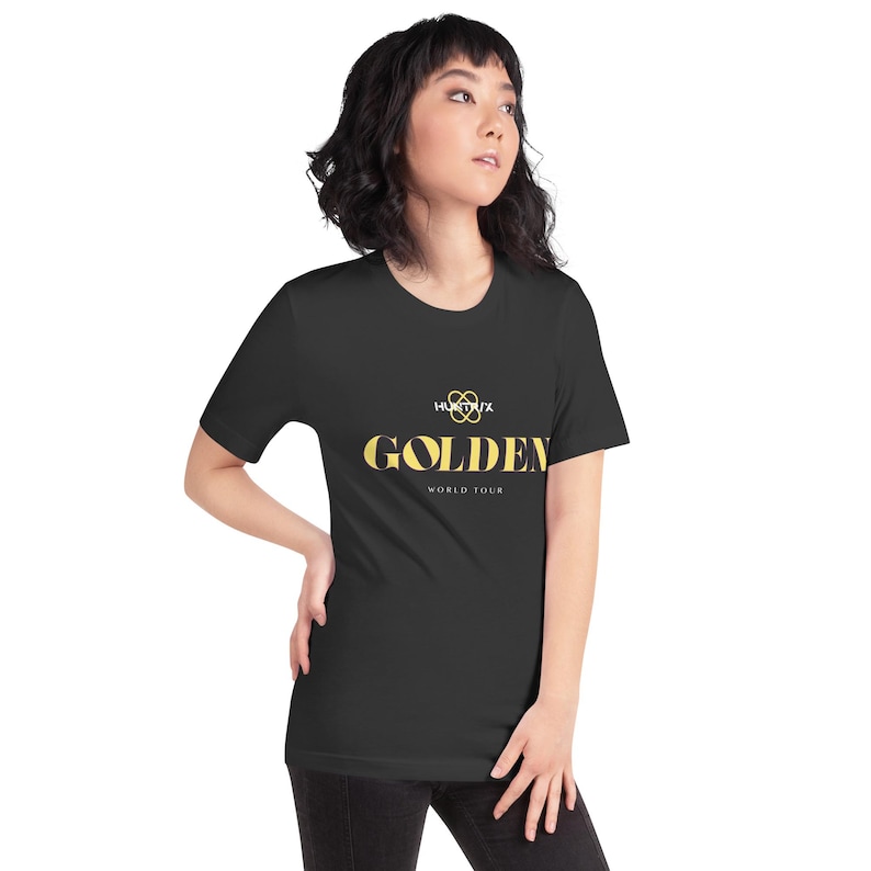 May include: Black t-shirt featuring the word "GOLDEN" in gold lettering. Above "GOLDEN" is a gold infinity symbol and the word "HUXTRUX". Below "GOLDEN" is "WORLD TOUR". The t-shirt is worn by a person.