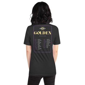 May include: Black t-shirt with the words "GOLDEN WORLD TOUR" in gold, and a list of tour dates and locations within a purple frame. The shirt has a crew neck and short sleeves.
