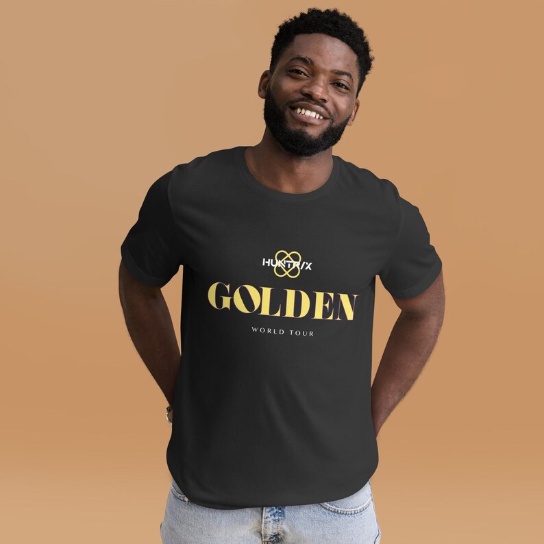May include: Black t-shirt featuring the word "GOLDEN" in yellow, with the text "HUNTX/X" and "WORLD TOUR". The shirt has a crew neck and short sleeves. The background is a plain tan colour.