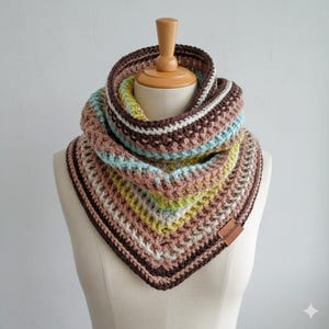 Peut inclure: Un snood crocheté à la main, multicolore. L'écharpe présente un mélange de rayures marron, roses, bleues et jaunes. Le snood est présenté sur un mannequin et porte une petite étiquette en cuir avec l'inscription "handmade".