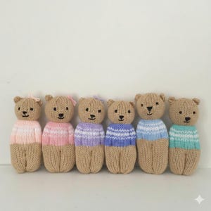 Peut inclure: Six ours en peluche tricotés à la main, alignés. Chaque ours a un corps beige et un pull rayé de différentes couleurs : rose, violet, bleu et turquoise. Certains ours ont un petit nœud. Les ours ont des yeux et des sourires brodés en noir.