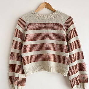 May include: A cream-coloured, striped jumper with long sleeves. It has horizontal stripes in a deep red colour. The neckline, cuffs, and hem are solid cream. The jumper is hanging on a wooden hanger against a white background.