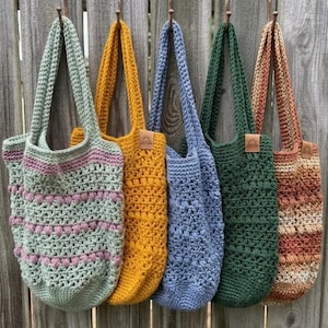 May include: Five handmade crochet tote bags hanging in a row. The bags feature various textures and colors, including sage green with pink accents, solid mustard yellow, solid periwinkle blue, solid forest green, and a multi-colored striped pattern.