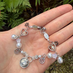 May include: A silver bracelet with a variety of charms, including heart-shaped pendants, faux pearls, small flower accents, and spiral designs. The bracelet is held in a hand against a backdrop of green foliage.