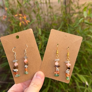 May include: Two pairs of dangle earrings featuring natural stone beads in green, orange, pink, brown, and grey. The earrings are presented on brown earring cards, set against a background of green foliage.
