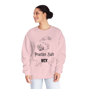 Puede incluir: Una sudadera rosa con un estampado gráfico negro de una mano apuntando a un libro con el texto "Practice Safe Hex" y un diseño de serpiente.