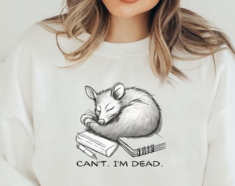 May include: White sweatshirt with a black and white illustration of a possum sleeping on top of books. The text "Can't. I'm dead." is printed below the possum.