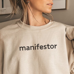 May include: A light beige sweatshirt with the word "manifestor" printed in black on the front.