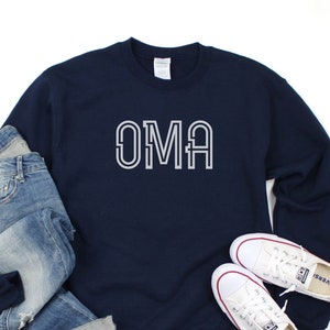 Peut inclure: Sweat-shirt bleu marine avec le mot "OMA" imprimé en blanc sur le devant. Le sweat-shirt est associé à un jean bleu et des baskets blanches.