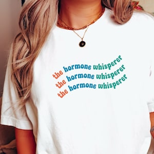 May include: White t-shirt with the text "the hormone whisperer" repeated three times in a rainbow-colored arc design.