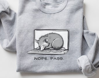 May include: A gray sweatshirt with a graphic of a possum sleeping inside a laptop. The text "NOPE. PASS." is printed below the graphic.