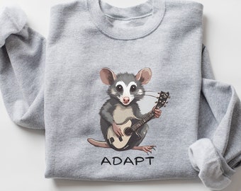 May include: A gray sweatshirt with a cartoon opossum playing a guitar. The opossum is wearing a white shirt and has a black eye patch. The text "ADAPT" is printed below the opossum.